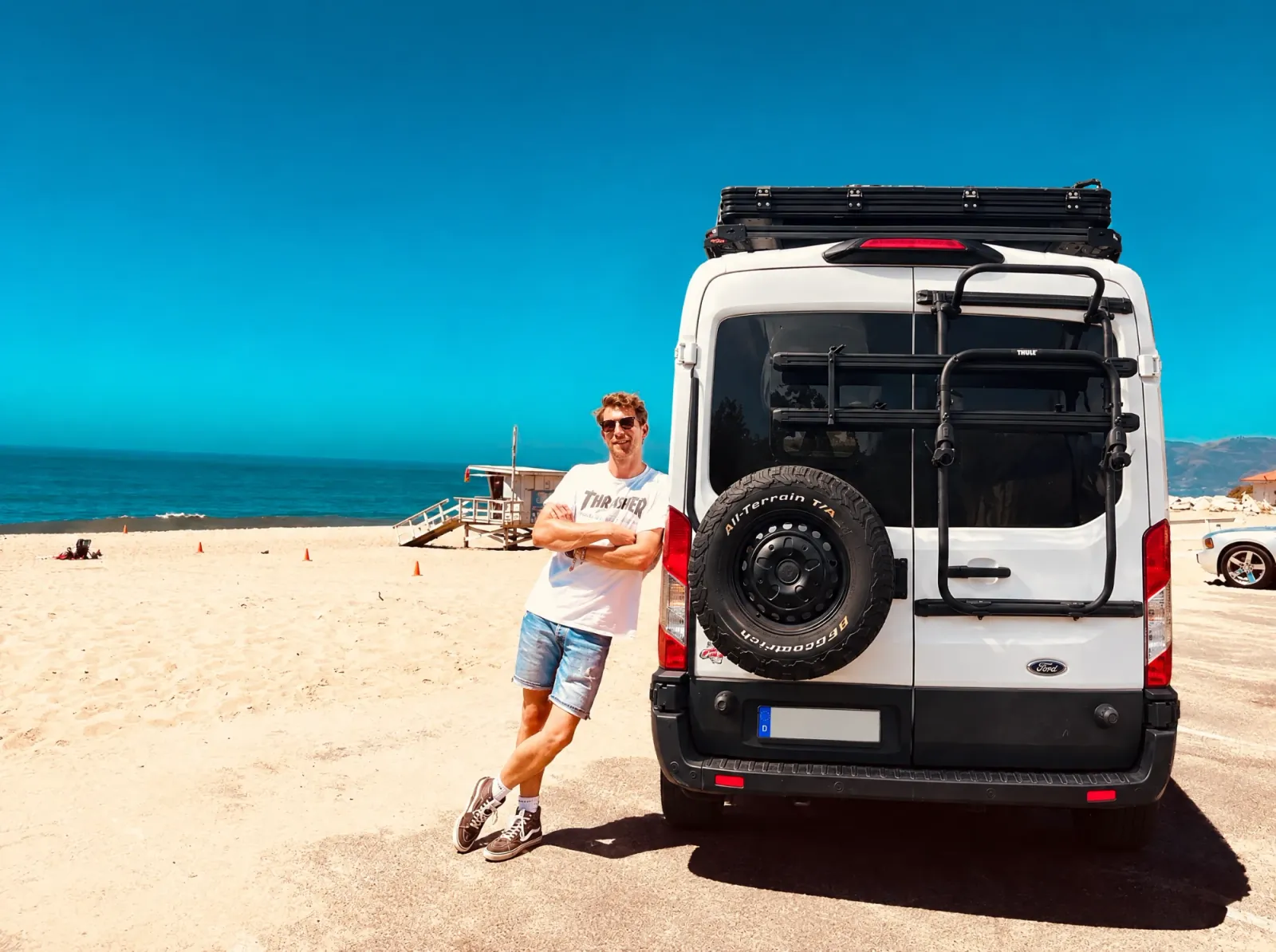 Oliver Tüngler, Gründer von 4RAMBLE, am Strand neben einem Ford Transit mit Dachträger und Reserverad